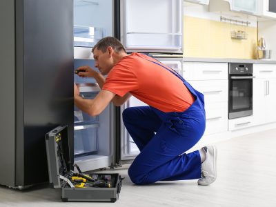 Male technician with screwdriver repairing refrigerator in kitchen
